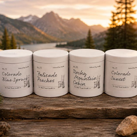 Four white metal tins of natural body care products lined up on a rustic wooden surface with rocks and evergreen branches, set against a scenic mountain lake at sunset.