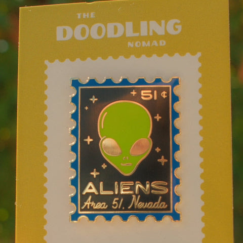 Enamel pin with alien design on a yellow card, held against a blurred green background.