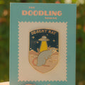 Desert Rat enamel pin on a card with 'The Doodling Nomad' branding, held against a blurred natural background.