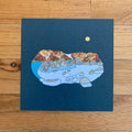 Illustration of a mountain landscape with a lake and moon on a blue card, placed on a wooden surface.