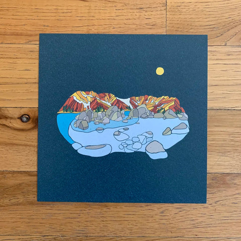 Illustration of a mountain landscape with a lake and moon on a blue card, placed on a wooden surface.