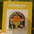 Enamel pin with alien in cowboy hat on a yellow card with 'The Doodling Nomad' branding.