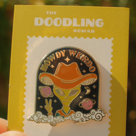 Enamel pin with alien in cowboy hat on a yellow card with 'The Doodling Nomad' branding.