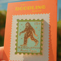 Enamel pin with Sasquatch design on an orange card, held against a blurred natural background.