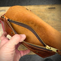 Brown leather pouch with a zipper held open by a hand on a wooden surface.