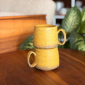 Two yellow ceramic mugs stacked on a wooden surface with a blurred indoor background.