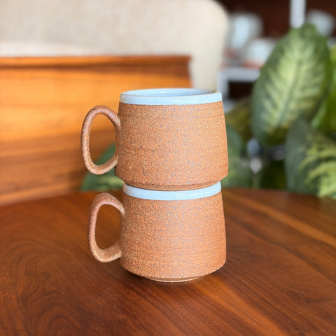 Two woven mugs stacked on a wooden surface with a blurred indoor background