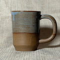 A handmade ceramic mug with a rustic two-tone glaze, featuring a textured light blue-gray upper half and a warm brown lower half, set against a neutral linen background.