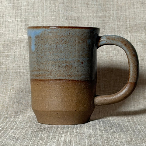 A handmade ceramic mug with a rustic two-tone glaze, featuring a textured light blue-gray upper half and a warm brown lower half, set against a neutral linen background.