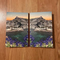 Two mountain landscape posters with blue flowers on a wooden surface