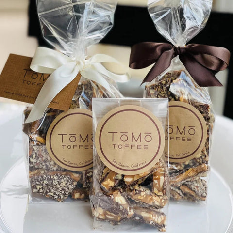 Three packages of Tomo Toffee with clear cellophane wrapping and ribbons on a white surface.