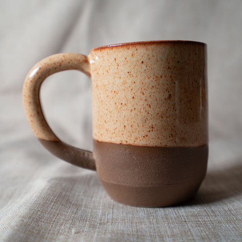 A handmade ceramic mug with a speckled beige and brown glaze and a solid dark brown base, set against a neutral draped fabric backdrop.