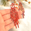Red circuit board design hanging ornament held in a hand with Christmas tree decorations in the background.
