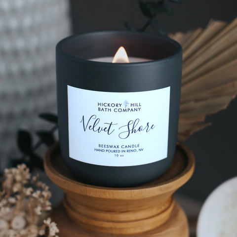 Black candle with a label on a wooden stand, surrounded by decorative elements.
