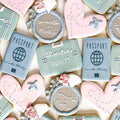Cookies shaped like suitcases, hearts, and passports with personalized inscriptions on a decorative background.