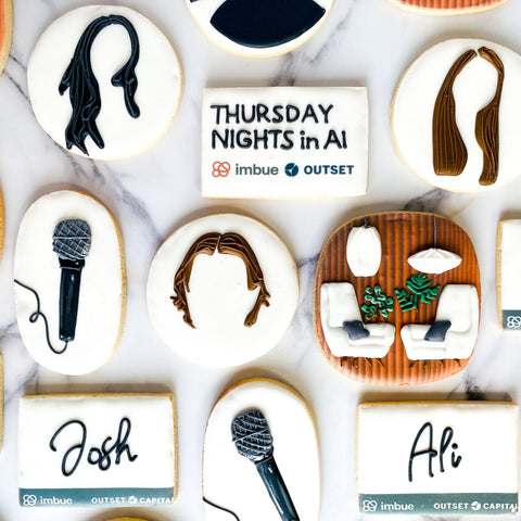 Decorative cookies with artistic depictions of people and objects on a marble surface, featuring event branding.