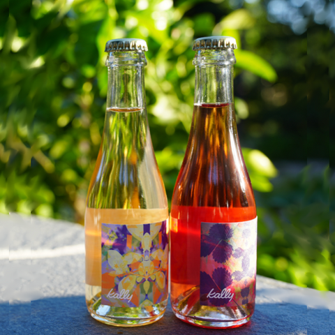 Two bottles of kally juice on a table with a blurred green outdoor background