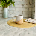 Ceramic mug on a woven placemat with a vase of flowers in the background.