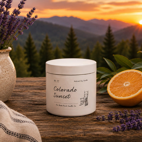 A white jar labeled 'Colorado Sunset' by Natural Tree Smells sits on a rustic wooden table, surrounded by lavender sprigs, sliced oranges, and a ceramic vase of lavender, with a mountain sunset landscape in the background.
