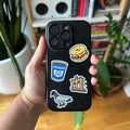 Person holding a black phone case with stickers in front of a bookshelf.