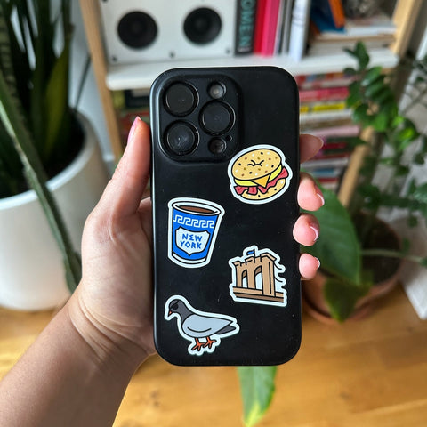Person holding a black phone case with stickers in front of a bookshelf.