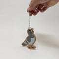 A hand holds a small ceramic bird ornament shaped like a pigeon, featuring a speckled gray and light blue body with a brown speckled base, hanging from a white string against a plain white background.