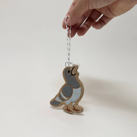 A hand holds a small ceramic bird ornament shaped like a pigeon, featuring a speckled gray and light blue body with a brown speckled base, hanging from a white string against a plain white background.