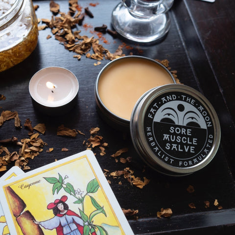Sore Muscle Salve container with candles and tarot cards on a dark surface