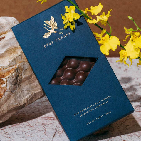 Blue chocolate box with a window displaying chocolate pieces, surrounded by yellow flowers on a textured surface.