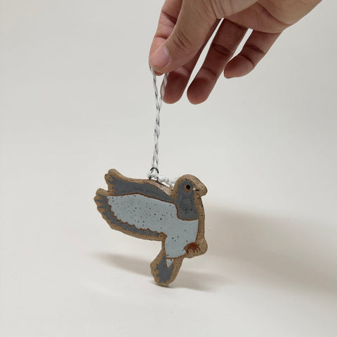 A hand holds a small bird-shaped ornament or charm with a silver string against a plain light background. The ornament has a pale blue-gray body with brown edges and darker gray wing details.