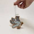 A hand holds a small bird-shaped hanging ornament against a plain light background. The ornament has a speckled light blue body, gray wings, and a tan speckled base, with a thin string loop at the top.