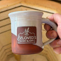 Person holding a mug with 'Beloved's Bakery & Cafe' logo on a wooden surface