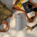 A matte light gray travel tumbler with a black lid sits on a white fabric surface, surrounded by travel props including a wooden tray with a passport, sunglasses, earbuds, keys, and a green bag.