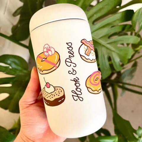 A hand holding a white insulated tumbler with a lid, decorated with cute donut illustrations and the words 'Hook & Press'. The background features large green monstera leaves.