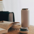 A matte off-white insulated can cooler with a metallic silver rim, shown with its lid and small dish on a woven table mat next to an open book.