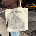 A person carrying a cream canvas tote bag with a black line art illustration of the New York City skyline and the text 'NEW YORK CITY' on the front, photographed on a city street with a yellow taxi in the background.