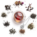 A clear glass teacup filled with amber tea sits on a matching saucer, surrounded by loose-leaf tea piles, dried flowers, sugar cubes, and a metal tea infuser on a white background.