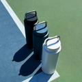 Three reusable insulated water bottles with metal handles are arranged diagonally on a green and blue outdoor court. The bottles are in black, dark blue, and light gray/silver, casting strong shadows on the court.