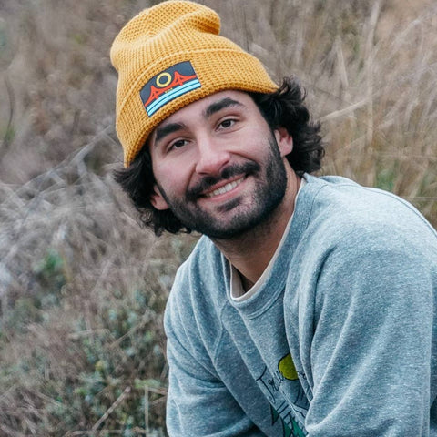 Man wearing a yellow beanie and light blue sweater sitting in a natural setting.