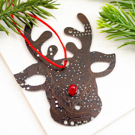 Reindeer-shaped ornament made from a circuit board with a red bead nose, surrounded by greenery on a white surface.