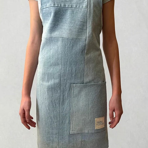 A light blue denim apron with a large front pocket and a small tan brand patch, shown on a model against a light gray background.