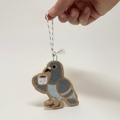 A hand holds a small ceramic bird ornament against a plain light background. The bird is light brown with gray wings and a pale blue belly, holding a small white cup.