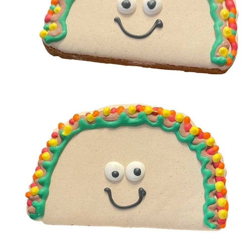 Two decorated sugar cookies shaped like tacos, with beige icing fillings, colorful candy-like border accents in red, orange, yellow, green, and pink, and simple face details on a white background.