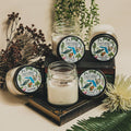 Candles in glass jars with decorative labels on a wooden stand, surrounded by plants.