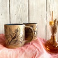Two ceramic mugs with floral designs on a pink surface, with a small vase in the background.