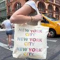 A woman on a city street carries a large cream canvas tote bag printed with 'NEW YORK CITY' repeated in pastel rainbow letters. A yellow taxi and brick buildings are visible in the background.