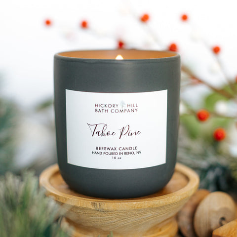 Candle labeled 'Tahoe Pine' from Hickory & Hill Bath Company on a wooden stand with a blurred background of greenery and berries.