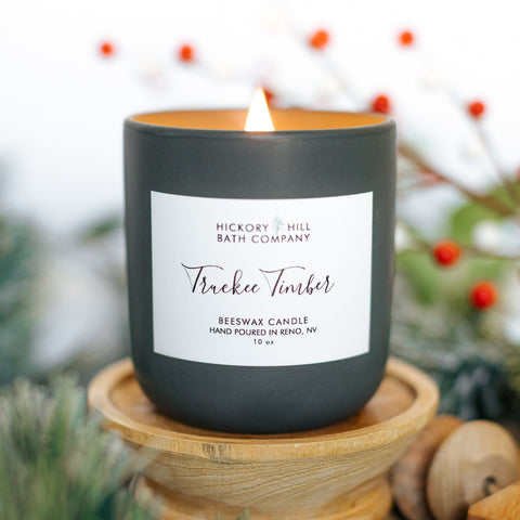 Black candle with a label on a wooden stand, surrounded by greenery and berries.