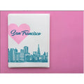 A white greeting card featuring a pink heart, the words 'San Francisco' in teal script, and a teal city skyline, photographed on a solid pink background.