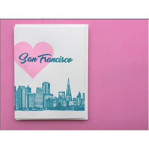 A white greeting card featuring a pink heart, the words 'San Francisco' in teal script, and a teal city skyline, photographed on a solid pink background.